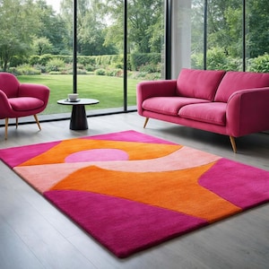 Puede incluir: Una vibrante y moderna alfombra con un diseño geométrico en tonos rosa, naranja y melocotón. La alfombra rectangular está colocada sobre un suelo de madera clara, complementando el sofá y las sillas rosas de la habitación.