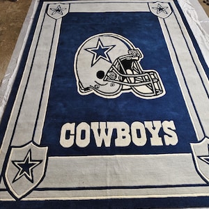 Puede incluir: Una alfombra rectangular con fondo azul oscuro y un gr&aacute;fico grande de un casco de f&uacute;tbol americano en blanco y plata en el centro. La palabra COWBOYS aparece en letras blancas debajo del casco, enmarcada por bordes plateados y estrellas.