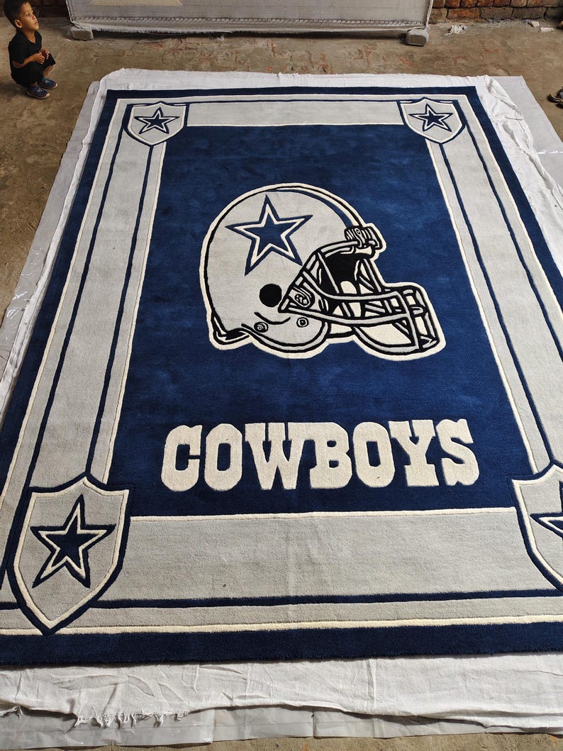 Puede incluir: Una alfombra rectangular con fondo azul oscuro y un gr&aacute;fico grande de un casco de f&uacute;tbol americano en blanco y plata en el centro. La palabra COWBOYS aparece en letras blancas debajo del casco, enmarcada por bordes plateados y estrellas.