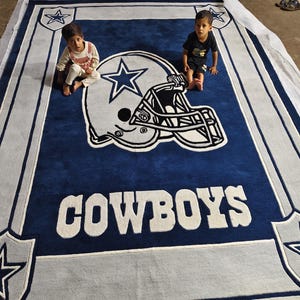 Puede incluir: Una alfombra rectangular grande con un fondo azul profundo y un gr&aacute;fico central de un casco de f&uacute;tbol americano en blanco y azul. La palabra COWBOYS aparece en letras blancas grandes debajo del casco, con logotipos de estrellas en las esquinas.