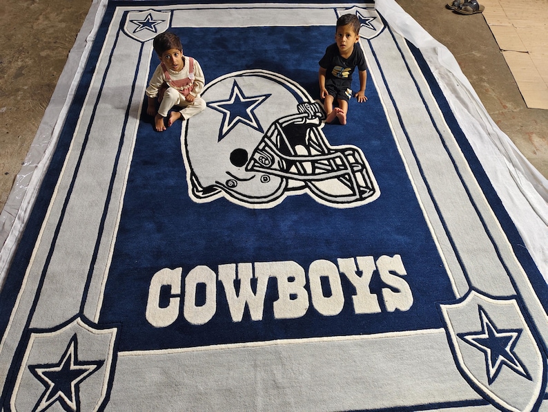 Puede incluir: Una alfombra rectangular grande con un fondo azul profundo y un gr&aacute;fico central de un casco de f&uacute;tbol americano en blanco y azul. La palabra COWBOYS aparece en letras blancas grandes debajo del casco, con logotipos de estrellas en las esquinas.