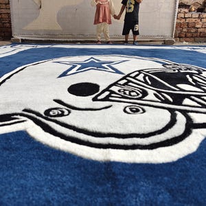 Puede incluir: Una alfombra grande de color azul, blanco y negro que presenta el logotipo de un casco de f&uacute;tbol americano de los Dallas Cowboys con una estrella central. La alfombra se muestra en el suelo con un dise&ntilde;o detallado de mechones y colores.
