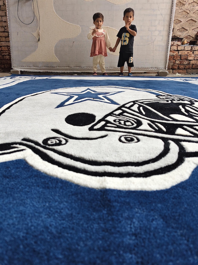 Puede incluir: Una alfombra grande de color azul, blanco y negro que presenta el logotipo de un casco de f&uacute;tbol americano de los Dallas Cowboys con una estrella central. La alfombra se muestra en el suelo con un dise&ntilde;o detallado de mechones y colores.