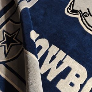 Puede incluir: Una alfombra de felpa con un esquema de colores azul profundo y gris claro. El dise&ntilde;o incluye un gran emblema de estrella, un gr&aacute;fico parcial de un casco de f&uacute;tbol americano y el texto "COWBOYS" en letras de bloque claras y negritas.