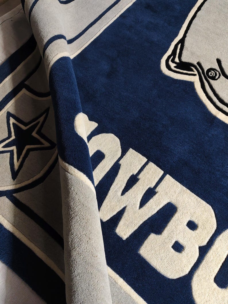Puede incluir: Una alfombra de felpa con un esquema de colores azul profundo y gris claro. El dise&ntilde;o incluye un gran emblema de estrella, un gr&aacute;fico parcial de un casco de f&uacute;tbol americano y el texto "COWBOYS" en letras de bloque claras y negritas.