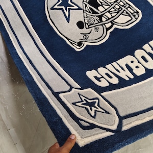 Puede incluir: Una alfombra rectangular de pelo insertado en azul oscuro, blanco y gris plateado. El dise&ntilde;o presenta un casco de f&uacute;tbol americano con un logotipo de estrella, el texto COWBOYS y un emblema de escudo con una estrella.