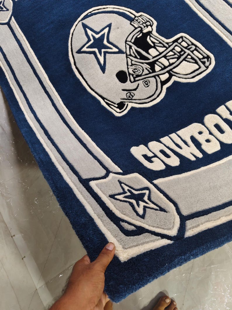 Puede incluir: Una alfombra rectangular de pelo insertado en azul oscuro, blanco y gris plateado. El dise&ntilde;o presenta un casco de f&uacute;tbol americano con un logotipo de estrella, el texto COWBOYS y un emblema de escudo con una estrella.