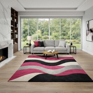 Puede incluir: Salón moderno con un sofá gris, una mesa de centro de madera y una alfombra colorida. La alfombra tiene un diseño ondulado en negro, rosa, gris y blanco. También se ven una chimenea y grandes ventanales con vistas a un jardín.