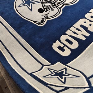 Puede incluir: Una alfombra rectangular con fondo azul profundo y un gr&aacute;fico grande de un casco de f&uacute;tbol americano en blanco y azul. La palabra COWBOYS aparece en letras blancas grandes a un lado, y un emblema de escudo con una estrella azul en la esquina.