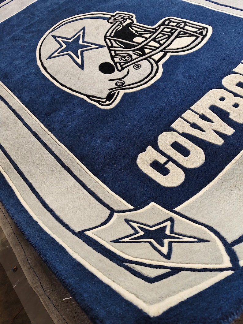 Puede incluir: Una alfombra rectangular con fondo azul profundo y un gr&aacute;fico grande de un casco de f&uacute;tbol americano en blanco y azul. La palabra COWBOYS aparece en letras blancas grandes a un lado, y un emblema de escudo con una estrella azul en la esquina.
