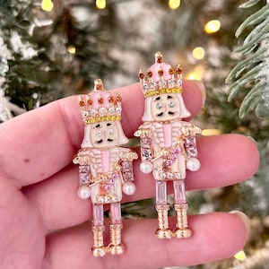 May include: Two decorative nutcracker figurines. Each figurine is white, pink, and gold, with a crown and gem accents. The nutcrackers are held in a hand against a blurred background of Christmas lights and greenery.
