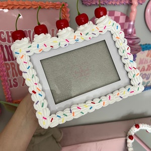 May include: White picture frame decorated to look like a cake with white frosting and colorful sprinkles. There are five red cherries on top of the frame.