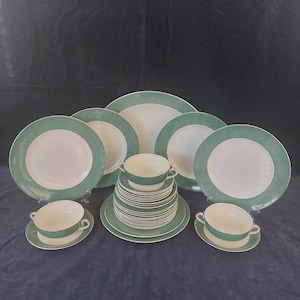 May include: A full dinner service with white plates and a green dotted edge. The set includes various plates, soup bowls, and saucers. The tableware is arranged on a dark blue surface.