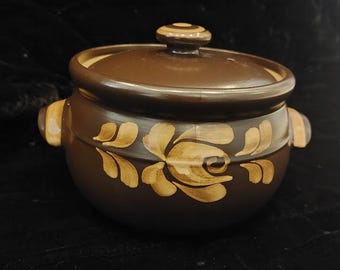 May include: A brown ceramic pot with a lid and two handles. The pot has a floral design painted in a lighter brown color.