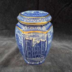 Op de afbeelding: Een blauw-witte keramieken pot met bijpassend deksel, voorzien van gedetailleerde illustraties van de kathedraal van Ripon. De pot is versierd met ingewikkelde bloemmotieven en een goudkleurige rand langs de opening en het deksel.