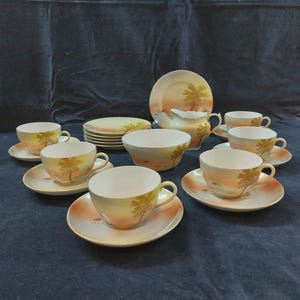 Puede incluir: Un juego de té vintage con tazas, platillos, platos, una crema y un cuenco. El juego tiene una base color crema con un diseño de paisaje en tonos naranja, amarillo y marrón. El diseño incluye un árbol y una puesta de sol.