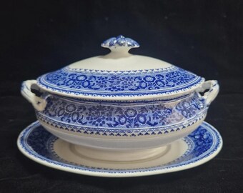 May include: A blue and white ceramic soup tureen with a matching lid and a saucer. The tureen has handles and a floral pattern. The lid has a decorative finial. The set is on a black surface.