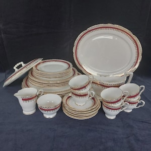 May include: A complete vintage porcelain dinner service. The set includes plates, cups, saucers, a serving platter, a gravy boat, a creamer, and a lidded dish. The pieces are white with a red and gold decorative border.