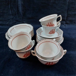 May include: A collection of white porcelain dishware with a red nautical design. The set includes cups, saucers, and bowls, each with a red rope-like border. The cups and bowls feature a red illustration of a sailing ship.