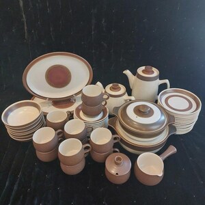 May include: A complete vintage stoneware dinnerware set in shades of brown and cream. The set includes plates, cups, saucers, a teapot, a serving platter, a covered casserole dish, and a creamer. The pieces have a circular design in the center.