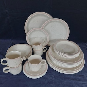 May include: A full dinner service set with plates, bowls, saucers, and mugs. The white ceramic items have a speckled design and a pink trim. The set includes plates and bowls of different sizes, plus several mugs and saucers.