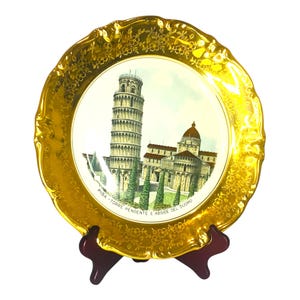 May include: A gold-rimmed ceramic plate with a depiction of the Leaning Tower of Pisa and the Duomo in Pisa, Italy. The plate is on a wooden stand. The text on the plate reads "PISA - TORRE PENDENTE E ABISIDE DEL DUOMO".