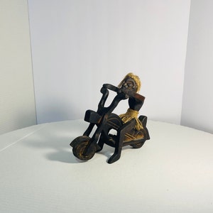 Puede incluir: Figurilla de madera de una persona montando una motocicleta. La figurita está tallada en madera oscura y tiene un diseño simple. La motocicleta tiene una sola rueda en la parte delantera y dos ruedas en la parte trasera.