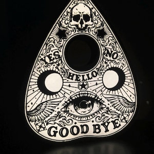 May include: A white planchette with black ink illustrations featuring a skull at the top, a central eye, and wings. The text includes YES, NO, HELLO, and GOOD BYE. Three circular cutouts are present, with ornate scrollwork surrounding the graphics.