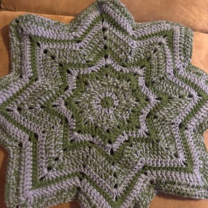 May include: A star-shaped crochet piece featuring alternating rows of sage green and light gray yarn. The design consists of a central circular pattern that expands into eight distinct points. An orange crochet hook rests above the textile.
