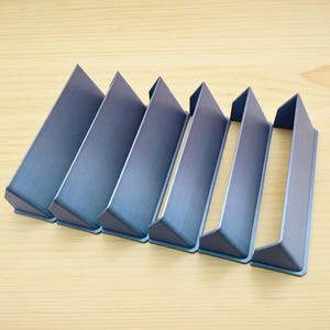 May include: A set of six blue metal drawer pulls with a brushed finish. Each pull features an angular, L-shaped profile with a flat front face and a triangular side support, designed for mounting onto the edge of a cabinet door or drawer front.