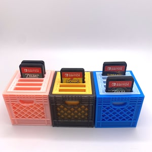 May include: Three mini crates, each 1.5 inches by 1.5 inches by 1.5 inches, are shown. The crates are made of plastic and are painted in different colors: pink, black, and blue. Each crate has five slots for Nintendo Switch game cartridges. The cartridges are visible in the slots, and the game titles are visible on the cartridges: Zelda, Sonic, and Human Fall Flat.