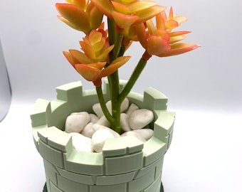May include: A light green ceramic planter shaped like a castle tower with a green base. The planter is filled with white stones and a small succulent plant with orange and yellow leaves.