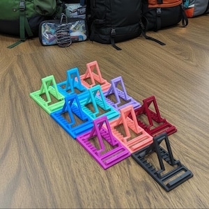 May include: A collection of ten adjustable plastic phone stands arranged in a grid. The stands are available in various colors including lime green, bright blue, light blue, pink, lavender, coral, dark red, magenta, and black, each with a notched base.