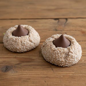 May include: Two peanut butter cookies with a chocolate kiss center. The cookies are light brown with a textured, speckled surface. The chocolate kisses are dark brown and cone-shaped. The cookies are on a wooden surface.