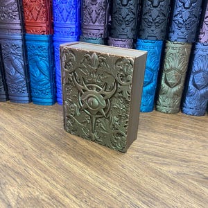 May include: A rectangular bronze-colored storage box featuring an embossed eye symbol at the center, surrounded by intricate floral scrollwork. The box has a hinged lid and is positioned in front of several other similar boxes in various colors.