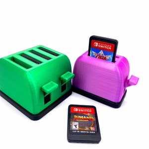 May include: Two 3D printed Nintendo Switch game cartridge holders shaped like toasters. One is green and the other is purple. A Nintendo Switch game cartridge is in the purple toaster. The game cartridge is for the game "The Legend of Zelda: Breath of the Wild".
