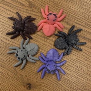 May include: Five colorful spider figurines in shades of brown, pink, black, gray, and purple. Each spider has eight legs and a detailed body. The spiders are arranged on a wooden surface.