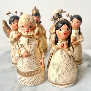 Pode incluir: Uma coleção de seis figuras de cerâmica vintage. As figuras estão vestidas com túnicas creme com detalhes dourados. As figuras têm rostos pintados com lábios vermelhos e cabelo preto. Algumas figuras têm asas ou adereços de cabeça dourados. As figuras provavelmente fazem parte de um presépio.