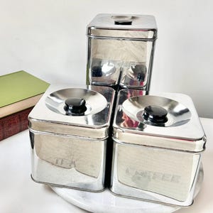 May include: A collection of three vintage chrome kitchen canisters. The canisters are labelled "FLOUR", "TEA", and "COFFEE" in embossed lettering. Each canister has a black knob on the lid. The canisters are square and have a shiny, reflective surface.