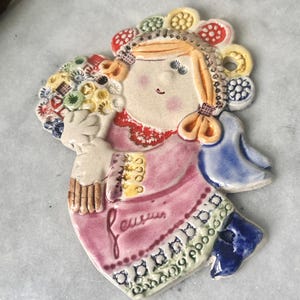 May include: A ceramic figurine of a girl holding a bouquet of flowers. The figurine is painted in shades of pink, blue, yellow, and white. The word "eusuis" is written on the dress. The girl has blonde hair and a decorative headpiece.