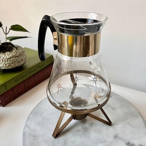 Puede incluir: Cafetera de cristal vintage con asa negra, detalles metálicos dorados y diseños de estrellas. La cafetera se apoya sobre un soporte de metal color latón. Un artículo de cocina clásico de mediados de siglo.
