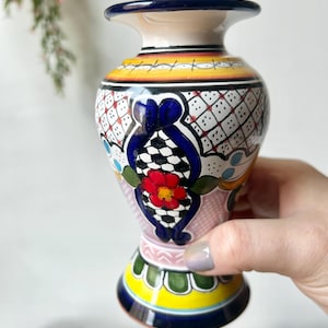 May include: A hand-painted ceramic vase with a flared rim and base. The vase features a vibrant design with a red flower, blue accents, and geometric patterns in black, white, and yellow. The vase is held by a person.