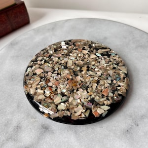 May include: A round, black coaster with a mosaic of small, iridescent shell fragments. The coaster has a glossy finish and a dark border. The shell pieces are in shades of beige, pink, and gold, creating a textured surface.