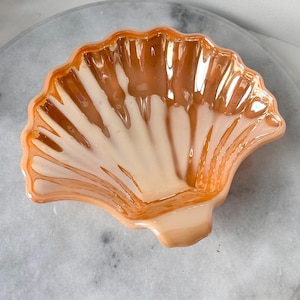 Puede incluir: Un plato de vidrio en forma de concha de vieira de color melocotón con un interior blanco iridiscente. El plato tiene una superficie lisa y brillante.