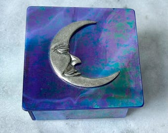 Può includere: Scatola portagioie quadrata con un design a mezzaluna argentata sul coperchio. La scatola ha una finitura marmorizzata viola e blu. La luna ha un volto dettagliato. È un oggetto decorativo per riporre oggetti.