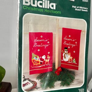 Könnte beinhalten: Ein Bucilla Christmas Heirloom-Kit für ein Paar Stickerei-Gästetücher. Die Verpackung zeigt zwei rote Handtücher mit gesticktem "Season's Greetings" und weihnachtlichen Designs. Das Kit enthält Stoff, Garn und eine Nadel.