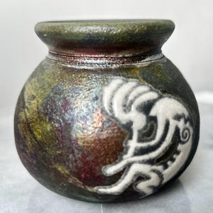May include: A small, round ceramic vase with a dark green and gold mottled finish. A white, stylized figure is painted on the side. The vase has a narrow neck and a wide, rounded body. The overall effect is rustic and artistic.