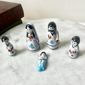 May include: A ceramic nativity set featuring five figurines. The figures are painted with white, blue, and orange details. The set includes Mary, Joseph, baby Jesus, and two other figures, all with black hair.