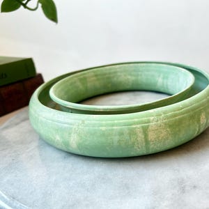 Peut inclure: Un bol en céramique vert clair de forme ronde avec un bord large et plat, contenant un bol intérieur plus petit. La glaçure présente une finition texturée et marbrée avec des stries crème plus claires sur toute la surface.