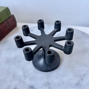 May include: A black metal candle holder with eight cylindrical candle cups arranged in a starburst pattern. The candle holder has a circular base and is designed to hold multiple candles. The finish is matte black.